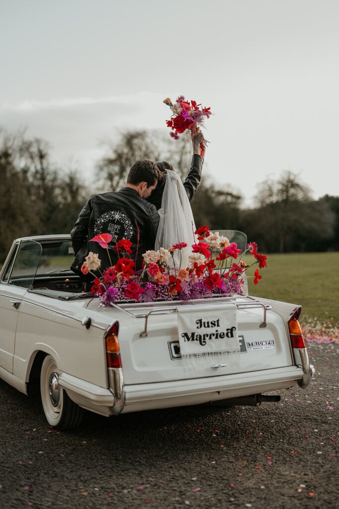 Image of the back of a white convertible with a wedding coupled sitting in the back amongst red and white flowers and a just married white banner hanging off the back of the car as they drive away. 