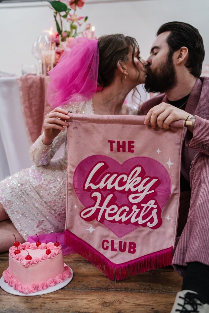 A couple kissing holding up a sign that says, "the lucky hearts club".