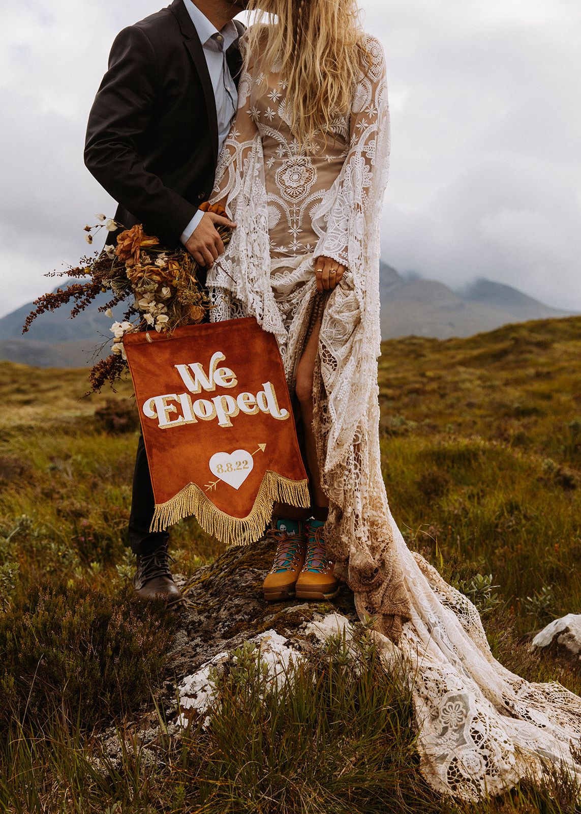 A couple standing on a grassy mountain side holding a, we eloped hand made sign by Daydreamer Creative Studio.
